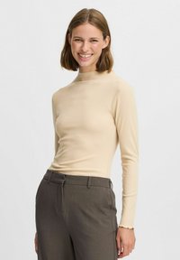 Beige geripptes Langarmoberteil mit Hohem Kragen und tailliertem Schnitt. Kombiniert mit dunkelgrauen Hosen mit Reißverschluss.