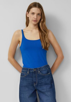 Młoda kobieta o długich włosach, ubrana w jasnoniebieską koszulkę na ramiączkach i ciemnoniebieskie jeansy z wysokim stanem, stoi na tle gładkiej jasnej ściany.