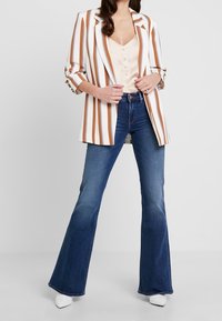 Randig blazer i vitt, brunt och svart, kombinerad med en beige topp och utsvängda blå denimjeans. Vita klackskor fullbordar outfiten.