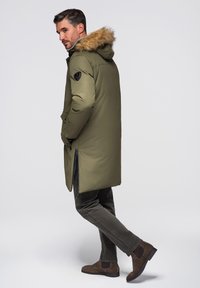 Mann trägt langen olivgrünen Wintermantel mit Pelzkapuze, dunkle Hose und braune Wildlederstiefel, posiert mit den Händen in den Taschen vor einfarbigem Hintergrund.