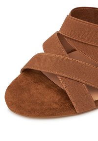 Sandalia de ante marrón con amplias tiras elásticas entrelazadas sobre la zona de los dedos y una suela lisa sobre un fondo blanco.