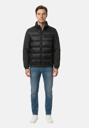 Uomo con giacca trapuntata nera, jeans blu e sneakers bianche in piedi davanti a uno sfondo chiaro uniforme.