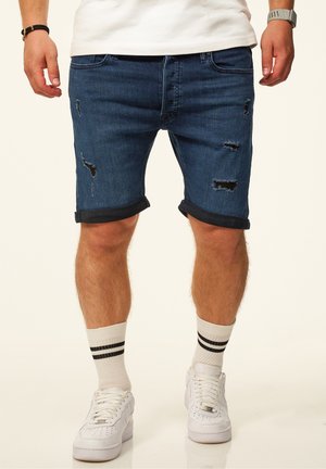 Mann trägt dunkelblaue hochgekrempelte, zerrissene Jeansshorts, weiße Sneaker und weiße Socken mit zwei schwarzen Streifen, steht vor einem schlichten Hintergrund.
