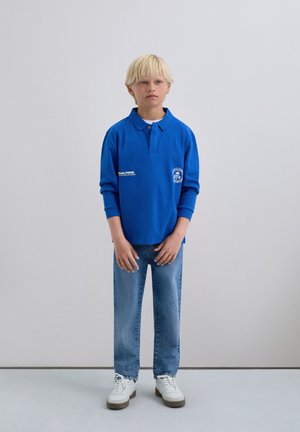 Camisa polo de manga larga azul con cuello y detalle de logo, combinada con jeans de pierna recta azul claro y zapatillas blancas con suelas grises.