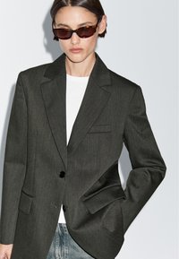 Blazer su misura grigio scuro con rever a punte, chiusura con un solo bottone e due tasche frontali, indossato sopra una camicia bianca.