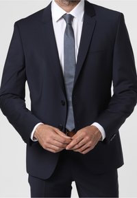 Námořnický modrý oblek s přiléhavým střihem, jednoradový design, dvě knoflíky a klopový límec. V kombinaci se světle modrou vzorovanou kravatou.