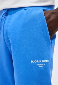 Κοντινό πλάνο χεριού σε τσέπη βασιλικά μπλε φόρμας με λευκό λογότυπο Björn Borg Stockholm 1956 στον μηρό, με λευκό φόντο.