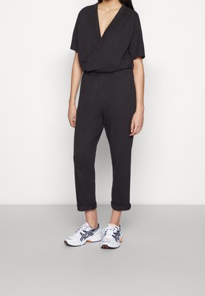 Kvinde iført en sort kortærmet jumpsuit med V-udskæring, op rullede ærmer og hvide atletiksko, står mod en ens hvid baggrund.