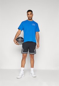 Camiseta de algodón azul con logo, pantalones cortos de malla negros con ribete blanco, sosteniendo un balón de baloncesto. Zapatillas deportivas blancas con detalles grises completan el look.