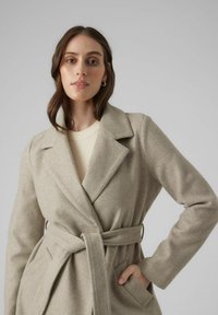 Lys beige indpakningsfrakke med reverskrave, bælte i taljen, sidelommer og blødt, tekstureret stof. Båret over en cremefarvet ribbestrikket sweater.