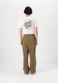 Weißes T-Shirt mit "SANTA CRUZ" Logo auf dem Rücken, kombiniert mit lockeren olivgrünen Jogginghosen, elastischem Bund und beigen Slide-Sandalen.