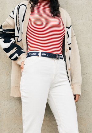 Osoba ubrana w czerwono-biały pasiasty top, beżowy kardigan z czarnymi wzorami, białe spodnie oraz czarny pasek z logo "Tommy Jeans".