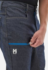 Jeans w ciemnoniebieskim odcieniu, z niebieską kieszenią zapinaną na zamek oraz białym logotypem "Millet" z przodu. Gładka tekstura i strukturalny kształt.