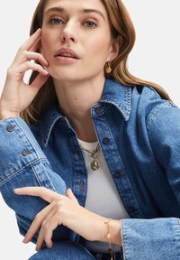 Helle blaue Denimjacke mit Druckknopfverschlüssen, über einem weißen, gerippten Oberteil getragen. Goldschmuck umfasst eine Halskette und ein Armband.