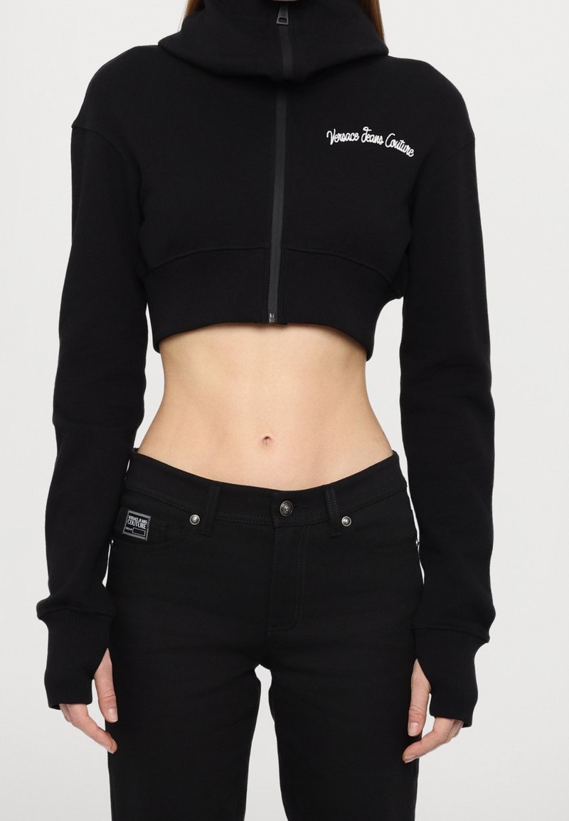 Sweat à capuche noir court avec col montant et fermeture éclair. Présente un logo brodé sur la poitrine. Associé à un jean en denim noir.