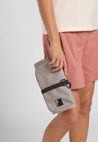 Grijze stoffen toilettas met een ritsluiting, zwarte strap en een merklabel, vastgehouden door een persoon in een roze korte broek.