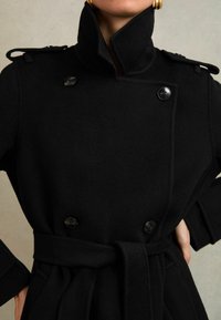 Melnas vilnas mētelis ar dubulto aizdari, lieliem melniem pogām, augstu apkakli un atbilstošu jostu. Iekļauj teksturētu audumu un strukturētas piedurknes.