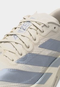 Κοντινό πλάνο αθλητικού παπουτσιού σε εκρού και γκρι με υφή από ύφασμα, δεμένα κορδόνια και εμφανές το όνομα της μάρκας στη γλώσσα.