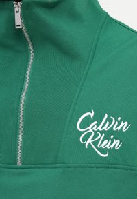 Zelen pulover na zadrgo z teksturiranim materialom, ki ima srebrno zadrgo in belo vezeno "Calvin Klein" logo na prsih.