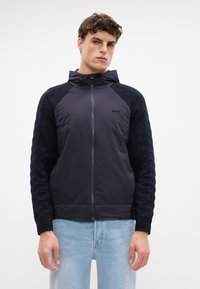 Casaco com capuz em azul marinho escuro, com um corpo de tecido liso e mangas em malha texturizada. Fecho frontal com zíper e detalhe do logótipo. Ajuste casual.