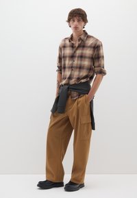 Camicia a quadri marrone con maniche arrotolate, abbinata a pantaloni cargo beige con tasche laterali e scarpe nere dalla suola spessa. T-shirt annodata in vita.