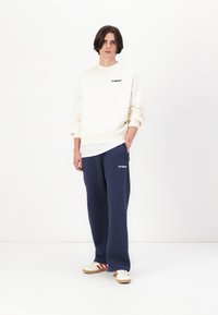 Cream sweatshirt with black logo, paired with navy sweatpants featuring the same logo. Casual white sneakers with red stripes and beige soles.