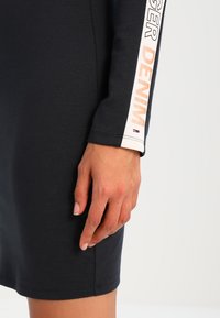 Main reposant à côté d'une robe noire à manches longues avec une bande blanche sur la manche affichant les mots "TOMMY JEANS DENIM."