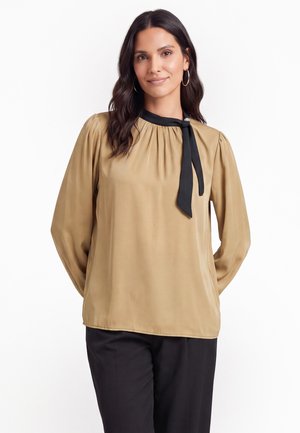 Vrouw met lang donker haar draagt een beige blouse met lange mouwen met een zwart dasspeldje en zwarte broek tegen een witte achtergrond.