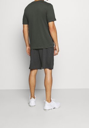Homme debout de dos portant une chemise à manches courtes vert foncé, un short gris au genou et des chaussures de sport blanches sur un fond uni.