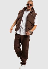 Brun polstret vest med hette over en hvit t-skjorte, sammen med brune cargo-bukser og hvite sneakers. Inkluderer synlige sømdetaljer.