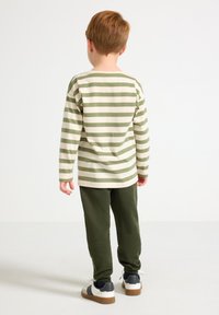 Ριγέ πουκάμισο με μακριά μανίκια σε κρεμ και ελιά με χαλαρή εφαρμογή, σε συνδυασμό με παντελόνι σε ελιά. Τα αθλητικά παπούτσια έχουν λευκές και ναυτικές λεπτομέρειες, καφέ σόλα.