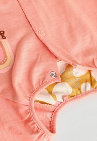 Ferskenfarvet baby-body med en cirkulær trykknaplukning ved skulderen. Foret med et mønstret stof med gule og hvide accenter.