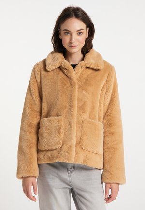 Jeune femme aux cheveux foncés portant une veste en fausse fourrure beige avec de grandes poches avant et un pantalon gris clair, regardant droit devant.