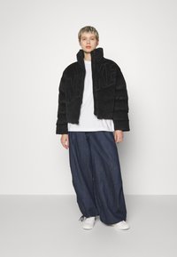 Svart kordjacka med stort "NIKE"-logotyp, ihop med löst sittande mörka denimbyxor och vita sneakers. Modellen står mot en enkel bakgrund.