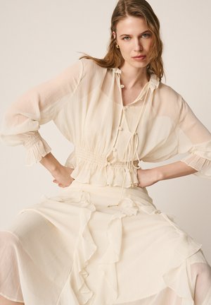 Kobieta ubrana w kremową, prześwitującą bluzkę z długim rękawem, z marszczonym taliowaniem, oraz pasującą, luźną spódnicę z falbankami, pozująca z rękami na biodrach.