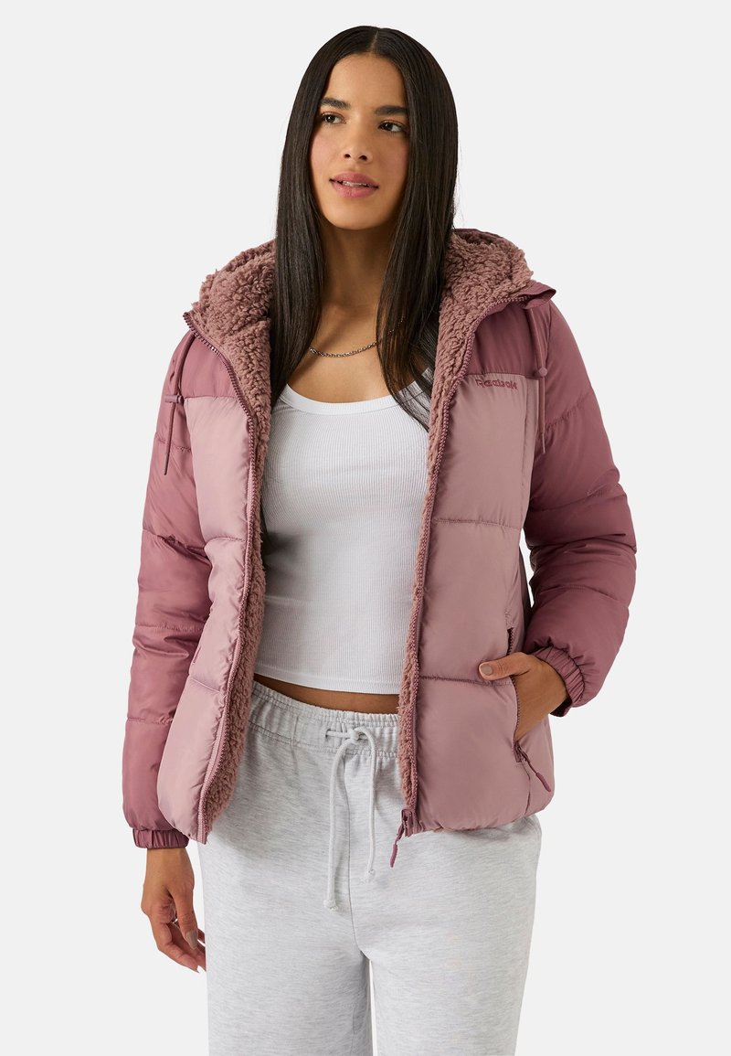 Różowa kurtka puchowa z miękkim kapturem podszytym kożuszkiem; pikowany wzór; długie rękawy; boczne kieszenie; w zestawieniu z białym topem i szarymi spodniami.