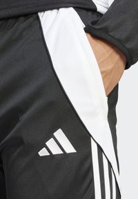 Svart-hvite treningstype bukser med elastisk liv, sidelommer, tre hvite striper og brodert logo på nedre del av benet.