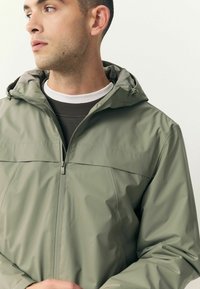 Jachetă ușoară, de culoare verde măsliniu, cu glugă, închidere frontală cu fermoar și o textură netedă, rezistentă la apă. Prezintă un design minimalist.
