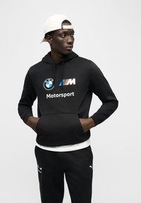 Mladý muž v černé mikině BMW Motorsport, černých kalhotách Puma a bílé kšiltovce obrácené dozadu, stojící s rukama v kapse mikiny.