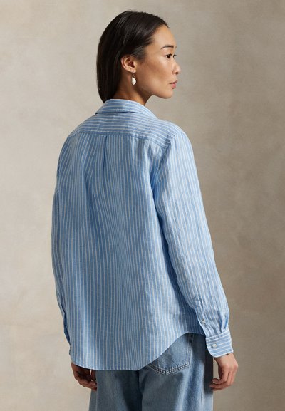 Femeie îmbrăcată într-un tricou cu mânecă lungă, cu dungi albastre și albe, și blugi albaștri, stând lateral pe un fundal bej simplu.