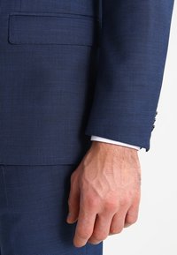 Abito blu navy in tessuto testurizzato con un motivo sottile. Caratterizzato da una tasca sul petto e polsini con dettagli di camicia bianca visibili.