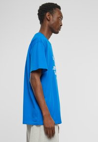 Blå bomulds t-shirt med korte ærmer og afslappet pasform; har hvid grafisk tekst på fronten; sidevisning, der viser minimale sømdetaljer.
