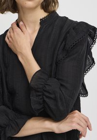 Blusa nera con un tessuto texturizzato, maniche arricciate con dettagli in pizzo e un collo alto. Linee sottili creano interesse visivo.