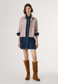 Giacca accorciata a righe beige e bianca sopra un vestito camicia in denim. La modella indossa stivali al ginocchio in suede marrone con tacco a blocco.