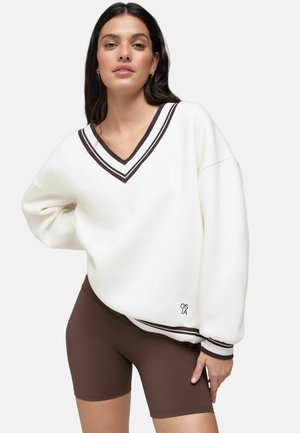 Vrouw draagt een oversized witte V-hals trui met zwarte strepen en bruine fietsbroek, staat met één hand op de heup.