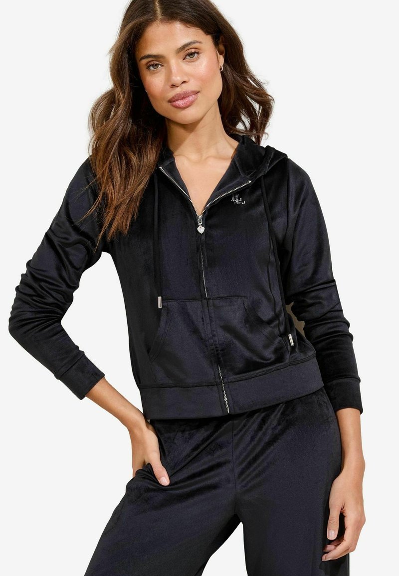 Crna velur zip-up dukserica s kapuljačom, prednjim džepom i srebrnim detaljima. Ima duge rukave i tijelo koje se dobro prilagođava.