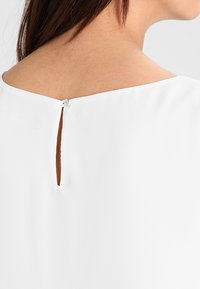 Weiße Bluse mit rundem Ausschnitt, einem kleinen Knopfverschluss auf der Rückseite und einem dezenten Schlitzdetail für ein elegantes Design.