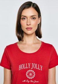 Červené bavlněné tričko s kulatým výstřihem, s bílým textem "HOLLY JOLLY" a kruhovým logem na přední straně.