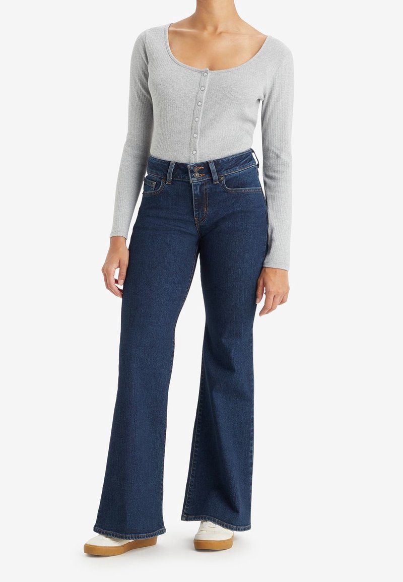 Femme portant un haut boutonné gris clair à manches longues, un jean évasé bleu foncé taille haute et des chaussures blanches avec des semelles beige, debout.