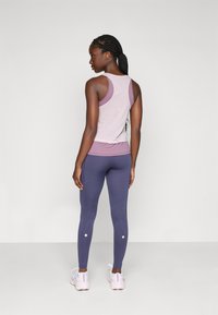 Woman with braided hair wearing a pink tank top, purple leggings, and white sneakers, standing and looking to the side against a plain background.